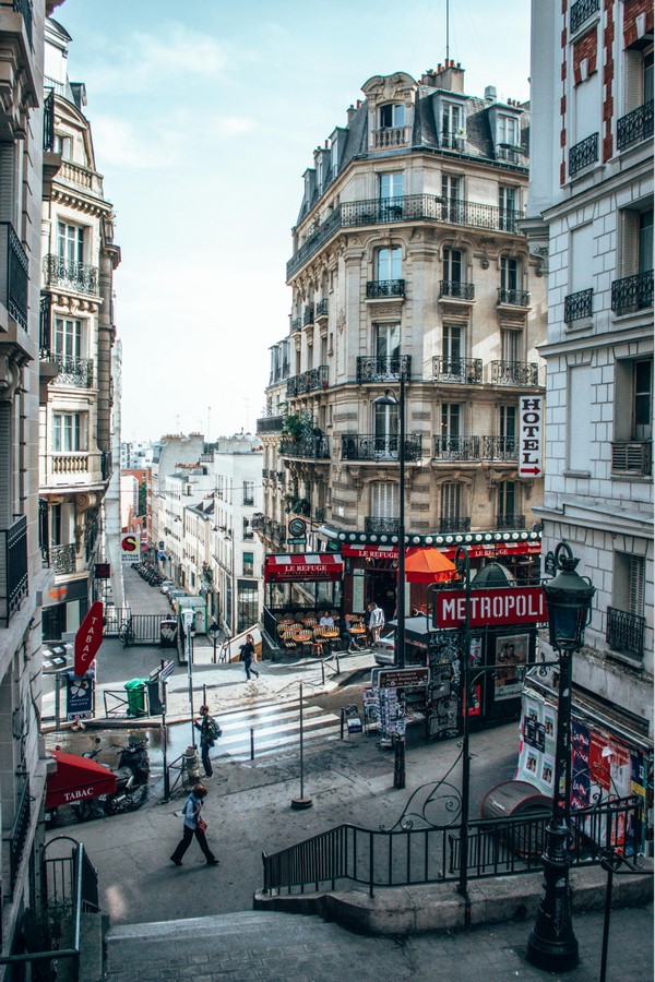 View of a Parisian street with traditional Haussmannian buildings featuring wrought iron balconies, a café with outdoor seating, and pedestrians walking by. The image includes a sign for 'Metropoli' and various storefronts.